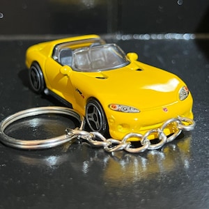 以下が含まれることがあります： 黄色いおもちゃの車のキーホルダー。 ミニチュアスポーツカーは黒い車輪と透明なフロントガラスを備えています。 キーホルダーはシルバーのチェーンとリングで取り付けられています。 車の側面には「Viper」の文字が印刷されています。