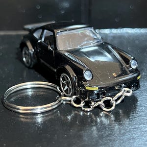 May include: A black Porsche sports car keyring featuring a die-cast metal body, a rear spoiler, clear windows, and silver-toned wheels. The vehicle is attached to a circular metal split ring by a short silver-toned chain connected to the front bumper.