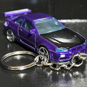 May include: A metallic purple toy car keychain featuring a black hood, a rear spoiler, and silver-toned wheels. The vehicle is attached to a circular metal split ring by a short silver chain, showcasing a detailed sports car design with clear windows.
