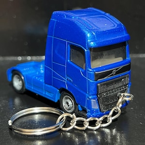 May include: A metallic blue Volvo semi-truck cab miniature attached to a silver metal keychain. The truck features a black front grille with the Volvo logo, clear windows, and silver wheels. A short metal chain connects the truck to a circular ring.