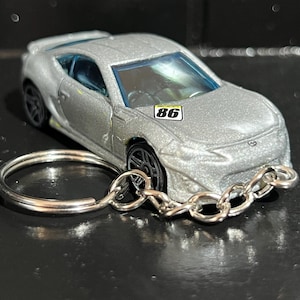May include: Silver toy car keyring with black wheels and blue tinted windows. The car has the number "86" on a small white and yellow sticker. The keyring has a silver ring and chain.