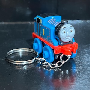 May include: A blue toy train with the number 1 on its side, red accents, and a smiling face. The train is attached to a silver keychain. The toy train is a popular children's character.