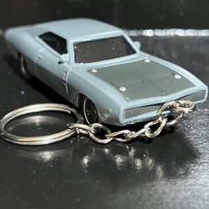 以下が含まれることがあります： ミニカーのキーホルダー。車は水色のクーペで、濃いグレーのボンネットが付いています。キーホルダーはシルバーの金属リングとチェーンが付いています。車は黒い窓とシルバーのホイールが付いています。