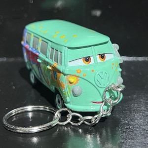 Op de afbeelding: Een turquoise speelgoedbusje met een sleutelhanger eraan. De bus heeft een cartoongezicht met bruine ogen en een rode glimlach. De bus is versierd met kleurrijke bloemen en abstracte ontwerpen.