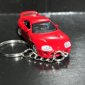 May include: A bright red toy car keychain featuring a detailed sports car model with a rear spoiler, clear windshield, and silver-colored wheels. A circular metal split ring is attached to the front of the vehicle by a short silver metal chain.