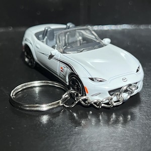 May include: A white Mazda MX-5 Miata convertible toy car keychain featuring black side stripes, a silver metal keyring, and a detailed interior with grey seats. The miniature vehicle has black wheels, clear headlights, and a visible Mazda logo on the hood.