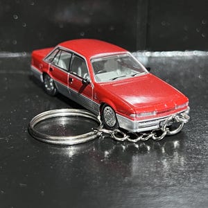 May include: A red and silver miniature car keyring. The toy car has a detailed design with silver rims and a chain attached to a silver keyring. The car is a classic sedan style.