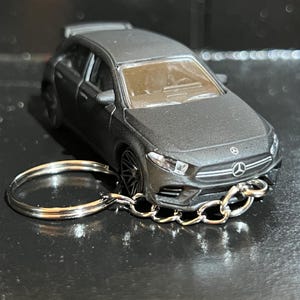 May include: A matte gray Mercedes-Benz A-Class model car keychain. The miniature car features detailed headlights, a front grill with the Mercedes logo, and a rear spoiler. The keychain has a silver chain and a key ring.