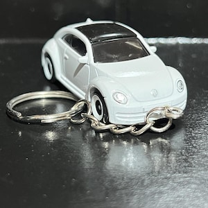 May include: A white toy car keyring with a black roof. The miniature vehicle has detailed features, including headlights and wheels. The keyring is attached to a silver metal ring and chain.