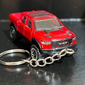 Puede incluir: Un camión de juguete rojo con ruedas negras y un llavero plateado. El camión tiene un diseño detallado, que incluye un techo corredizo y una parrilla delantera con la palabra "RAM". El llavero está sujeto a la parte delantera del camión.