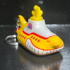 Op de afbeelding: Een gele onderzeeër sleutelhanger met een witte bovenkant en rode accenten. De onderzeeër heeft een lachend gezicht en een gele schoorsteen. De sleutelhanger is bevestigd aan een zilveren ring.