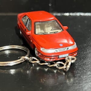 May include: A red miniature car keyring with a silver key ring and chain. The car has a detailed design, including windows, headlights, and a number plate. The keyring is on a dark surface.