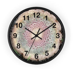 May include: A round wall clock with a black frame. The clock face features a detailed, textured design in shades of pink, green, and beige, resembling a cross-section of a tree trunk. Black hands indicate the time, and black numerals mark the hours.