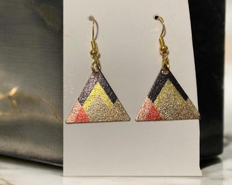 May include: A pair of gold triangle earrings with black, yellow, and pink geometric designs. The earrings have a glitter finish.