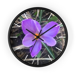 May include: A purple crocus flower with a yellow center is the focal point of this clock face. The flower is surrounded by brown and green foliage. The clock hands are black and the numbers are white.