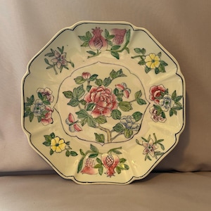Puede incluir: Un plato decorativo octogonal con fondo amarillo pálido y borde negro. El plato presenta un diseño floral central con flores rosas y rojas, rodeado de motivos florales y granadas más pequeños.