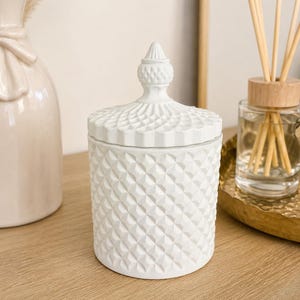 May include: A white cylindrical jar with a matching lid, featuring a textured diamond pattern across the exterior surface. The lid is topped with a pointed, decorative finial. The jar is opaque and matte white, showcasing a geometric design.