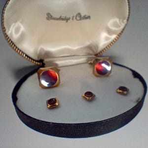 May include: A vintage set of cufflinks and shirt studs in a presentation box. The gold-toned cufflinks and studs have a square shape with a red and silver reflective center. The box is lined with white satin and has the text "Stradbridge & Co." printed on the inside.