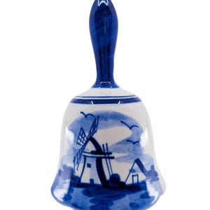 May include: A white ceramic bell with a blue handle and a blue hand-painted scene featuring a windmill, small buildings, and birds in flight. The bell has a flared base and a glossy finish, showcasing a classic blue and white color scheme.