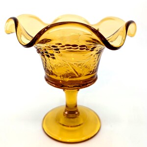 May include: Amber-colored glass pedestal bowl with a ruffled rim. The bowl features a detailed grape and leaf pattern around the sides. The base is round and the stem is slender, creating an elegant vintage aesthetic.