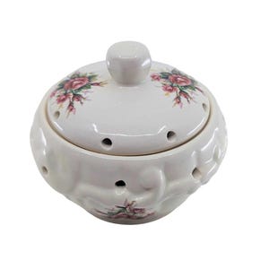 May include: A cream-colored ceramic pot with a matching lid, featuring a floral pattern of pink roses and green leaves. The round container has a series of small circular ventilation holes around the upper rim and a decorative embossed swirl design.