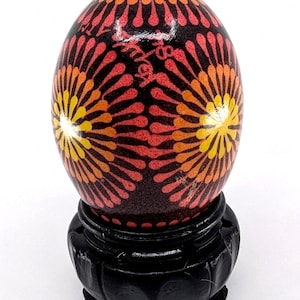 May include: A decorative egg with a black base, featuring a vibrant pattern of red, orange, and yellow floral designs. The egg is displayed on a black wooden stand, showcasing the intricate artwork.
