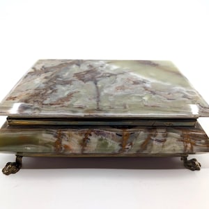 May include: A rectangular jewelry box with a marbled green and brown stone top and base. The box rests on four small, ornate brass feet. The box is a decorative storage item.