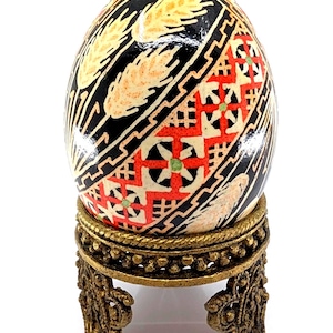 May include: A decorative egg with a black background and wheat stalks, geometric patterns in red, white, and green, and a gold-colored stand. The egg is an Easter or decorative item.