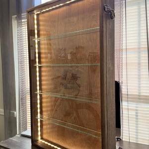 May include: A wooden display case with glass shelves and interior LED lighting. The case has a rectangular shape with a hinged door and metal clasps. The shelves are made of glass and are evenly spaced. The case is sitting on a dark brown desk.