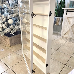 May include: A white display cabinet with a glass door, open to reveal several wooden shelves. The cabinet has black hardware and is standing on a tiled floor. The cabinet is next to a Christmas tree.