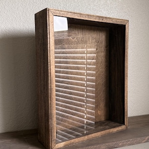 May include: A wooden shadow box with a clear glass front. The box is stained a medium brown and is rectangular. The interior is visible through the glass, with the shadow of blinds creating a striped pattern.