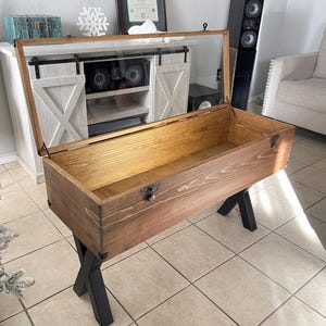May include: A wooden display case with a hinged glass top, resting on black metal legs. The case has a natural wood finish and is secured with metal latches. The design features an open interior, suitable for showcasing items.