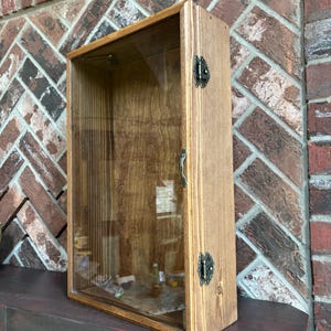 May include: A wooden display case with a glass front and a hinged door. The case is made of light-colored wood and has decorative metal hardware. The interior is visible through the glass, and the case is sitting on a dark surface.