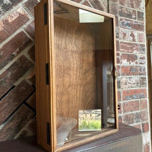 May include: A wooden display case with a glass front. The case is rectangular with a hinged door and black metal hardware. The wood has a warm, natural finish, and the interior is empty.