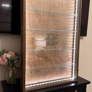 May include: Wooden display case with interior LED lighting. The case has a light brown wood finish, glass shelves, and a dark brown frame. The text "Bryan" is visible on the wood. The case is sitting on a black dresser.