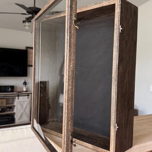 May include: A rectangular wooden display case with a dark brown finish and a hinged glass door. The interior features a black backing, and the frame is secured with small metal latches on the side. The case is open, revealing the empty interior.