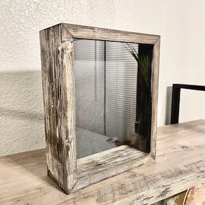 May include: A rustic, rectangular shadow box with a distressed, whitewashed wood frame. The box has a clear glass front and is displayed on a light-colored wooden surface. The frame's texture and color variations add to its vintage appeal.