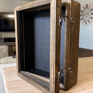 May include: A rectangular wooden shadow box display case with a dark brown stained finish. The frame features a hinged door with two antique brass-colored metal latches on the side and a black interior backing for displaying items securely.