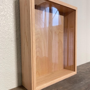 May include: A rectangular wooden display case with a clear glass front. The case is made of light-colored wood and is designed to be mounted on a wall. The interior is empty, showcasing the wood grain.