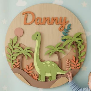 May include: A circular wooden wall decoration with a green dinosaur, palm trees, and the name "Danny". The artwork is on a light blue wall. A baby in a blue onesie is sitting on the floor.