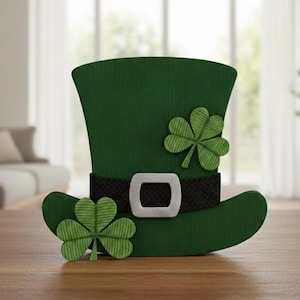 Op de afbeelding: Een groene leprechaun hoed voor St. Patrick's Day, met een zwarte riem en een zilveren gesp. Twee groene klavers zijn aan de hoed bevestigd. De hoed staat op een houten oppervlak.