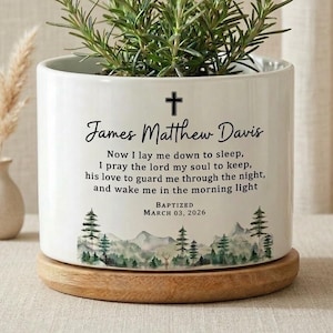 May include: A white ceramic planter with a cross and the name "James Matthew Davis" in script. The planter has a landscape design at the bottom and contains a green plant. The text reads "Now I lay me down to sleep... Baptized March 03, 2026."
