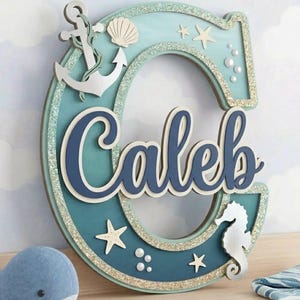 May include: A wooden letter "C" with the name "Caleb" in blue script, decorated with nautical elements like an anchor, seahorse, and starfish. The letter is teal with glitter accents, perfect for a nursery or child's room.