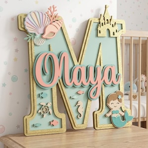 May include: A large, decorative wooden letter "M" in a sea-themed design. The letter is light blue with gold trim and features the name "Maya" in pink script. Adorned with a seashell, coral, a castle, and a mermaid, along with sea creatures.