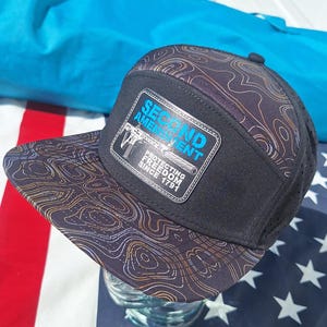 Peut inclure: Une casquette snapback noire à cinq panneaux avec un motif de carte topographique aux lignes orange et bleues sur la visière et la calotte. Un écusson rectangulaire affiche le texte SECOND AMENDMENT PROTECTING FREEDOM SINCE 1791 et un fusil.