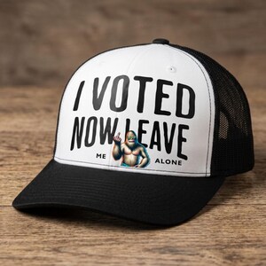 Op de afbeelding: Een zwart-witte truckerpet met een wit voorpaneel met de zwarte tekst I VOTED NOW LEAVE ME ALONE en een afbeelding van een bruin wezen met een zonnebril dat de middelvinger opsteekt. De pet heeft een zwarte klep en een mesh achterkant.