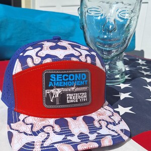 May include: A five-panel baseball cap featuring a blue and white topographic map pattern, a solid red front panel, and a rectangular patch. The patch displays the text SECOND AMENDMENT PROTECTING FREEDOM SINCE 1791 with a rifle graphic.