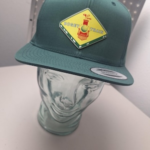 Könnte beinhalten: Eine tealfarbene Baseballkappe mit flachem Schirm und einem gelb-grünen Aufnäher mit der Aufschrift "Bogey Train". Der Aufnäher zeigt einen Golfball, Golfschläger und einen Zug. Die Kappe wird auf einem durchsichtigen Glaskopf präsentiert.