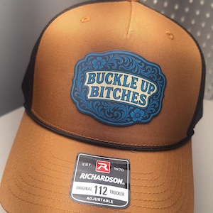 May include: A brown trucker hat with a black mesh back and a curved peak. The hat features a blue and white patch with the text "BUCKLE UP BITCHES". A Richardson 112 Trucker label is visible.