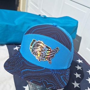 May include: A blue and black baseball cap featuring a topographic line pattern on the brim and crown. The front panel displays a holographic patch of an eagle silhouette over a map of the United States with the text 250 YEARS 1776-2026.